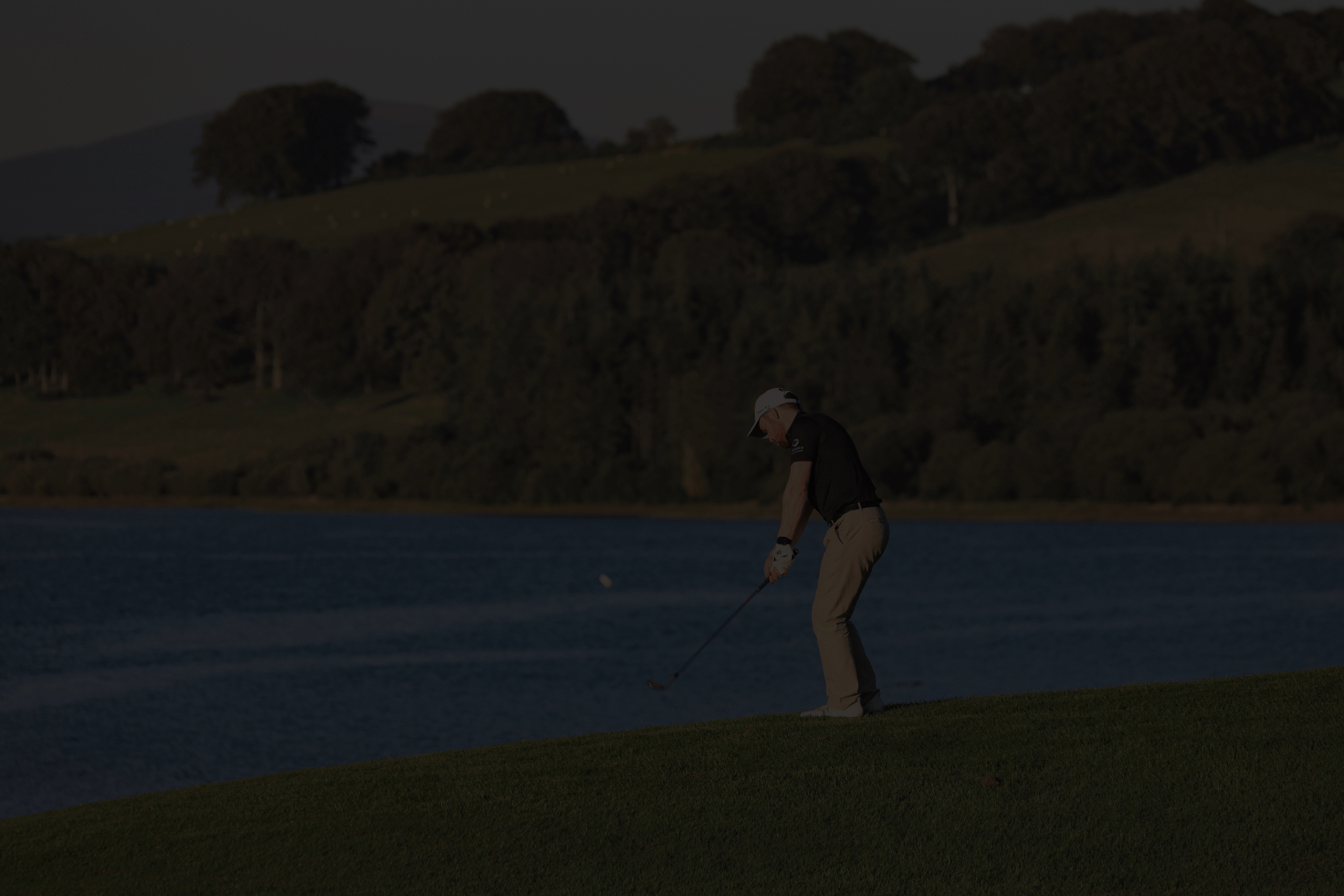 LGBT Golf in London. Gary Shaw Golf hits a wedge shot from the rough with a lake in the background.
